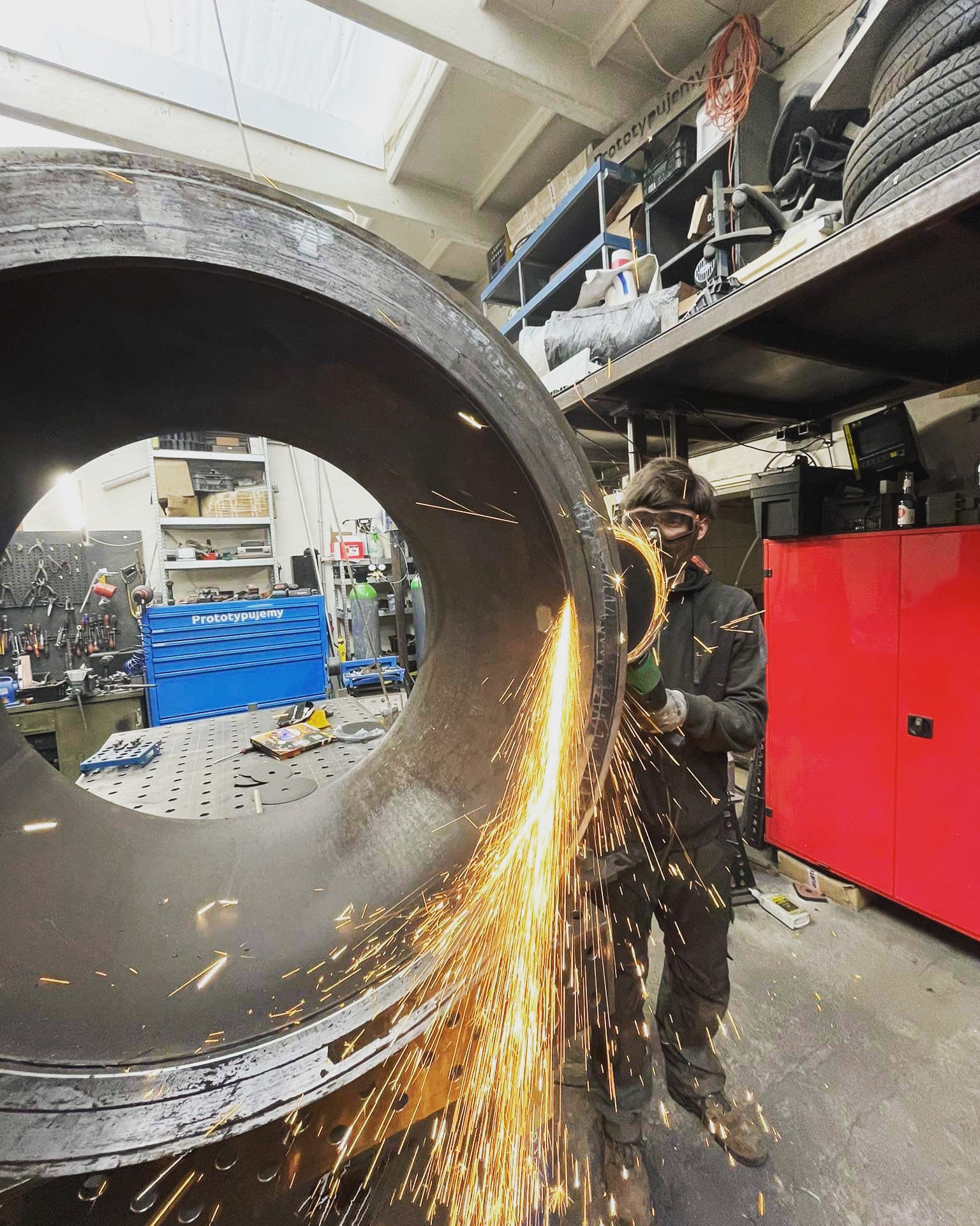 Pracownik w okularach ochronnych szlifuje spaw dużej, metalowej rury przy użyciu szlifierki kątowej, iskry rozbryzgują się w warsztacie.