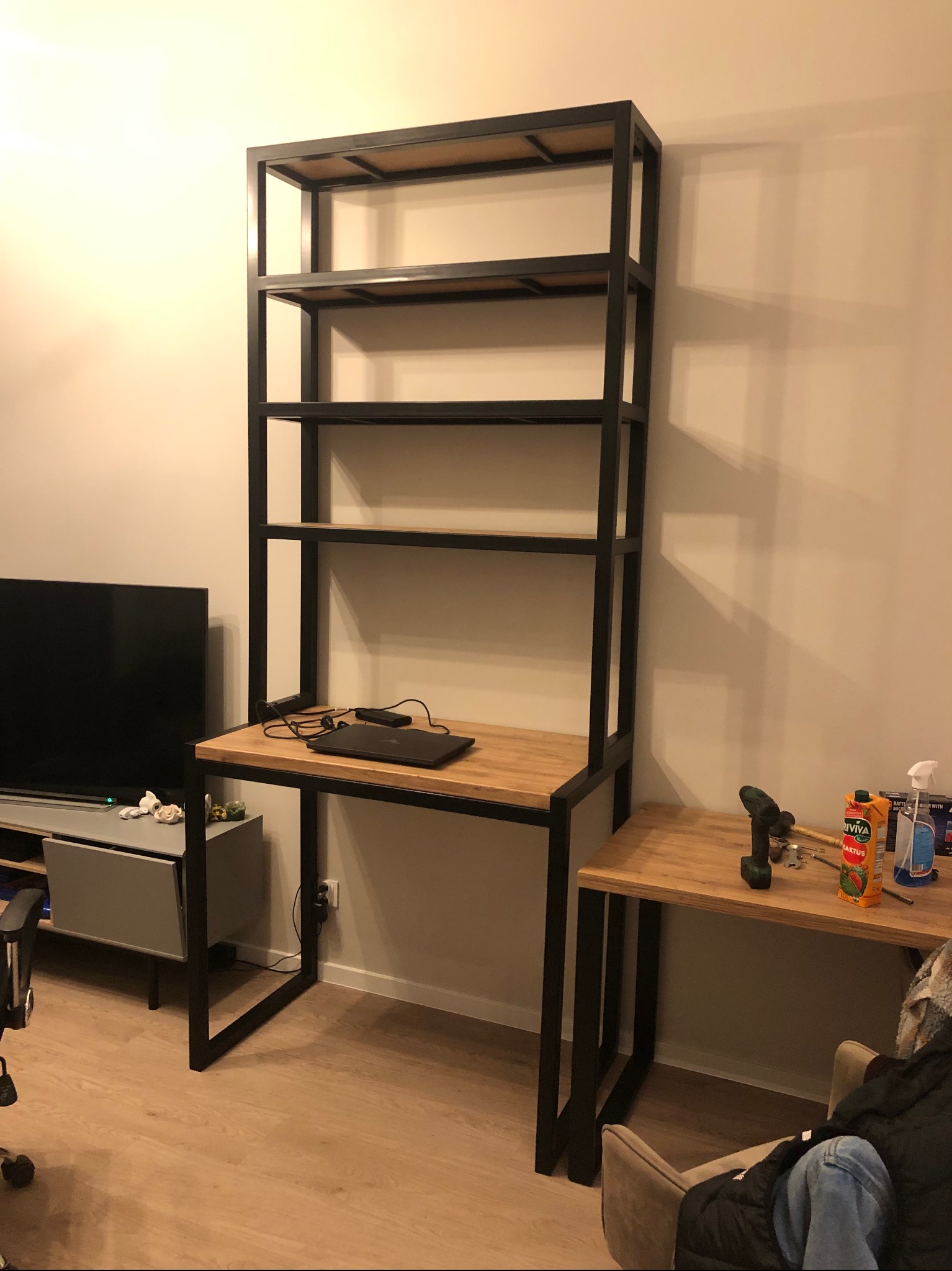 Metalowa konstrukcja regału z biurkiem z drewnianym blatem w nowoczesnym wnętrzu, widoczny laptop, narzędzia i napój.