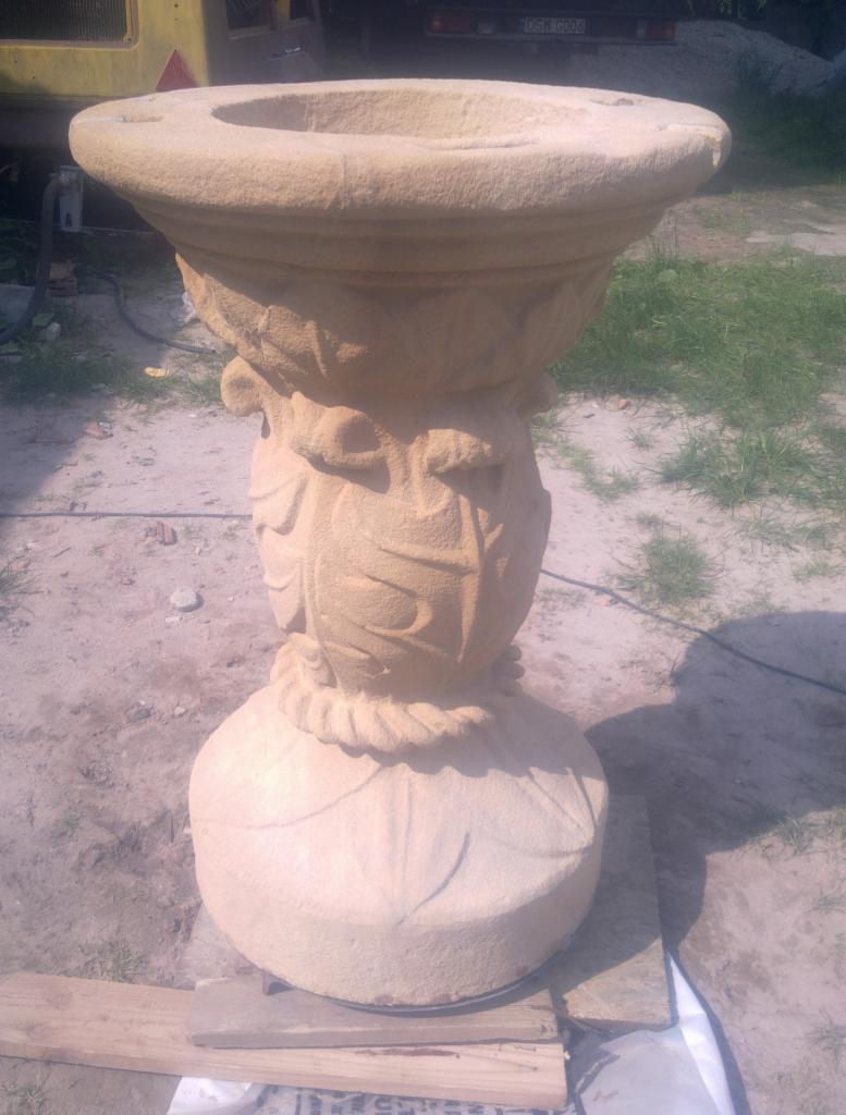 Piaskowcowa, zdobiona donica ogrodowa na postumencie, widoczna na zewnątrz, z elementami roślinnymi w rzeźbieniu.