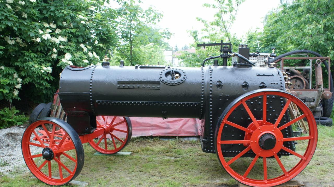 Czarny, nitowany parowóz z czerwonymi kołami na trawiastym terenie, z fragmentem innego urządzenia w tle.