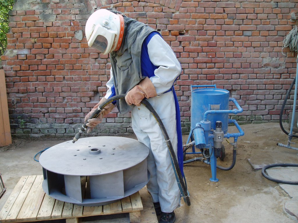 Pracownik w kombinezonie ochronnym i hełmie piaskuje metalową turbinę wentylacyjną na drewnianym podeście, widoczny niebieski zbiornik i węże do piaskowania, tło stanowi ceglana ściana.