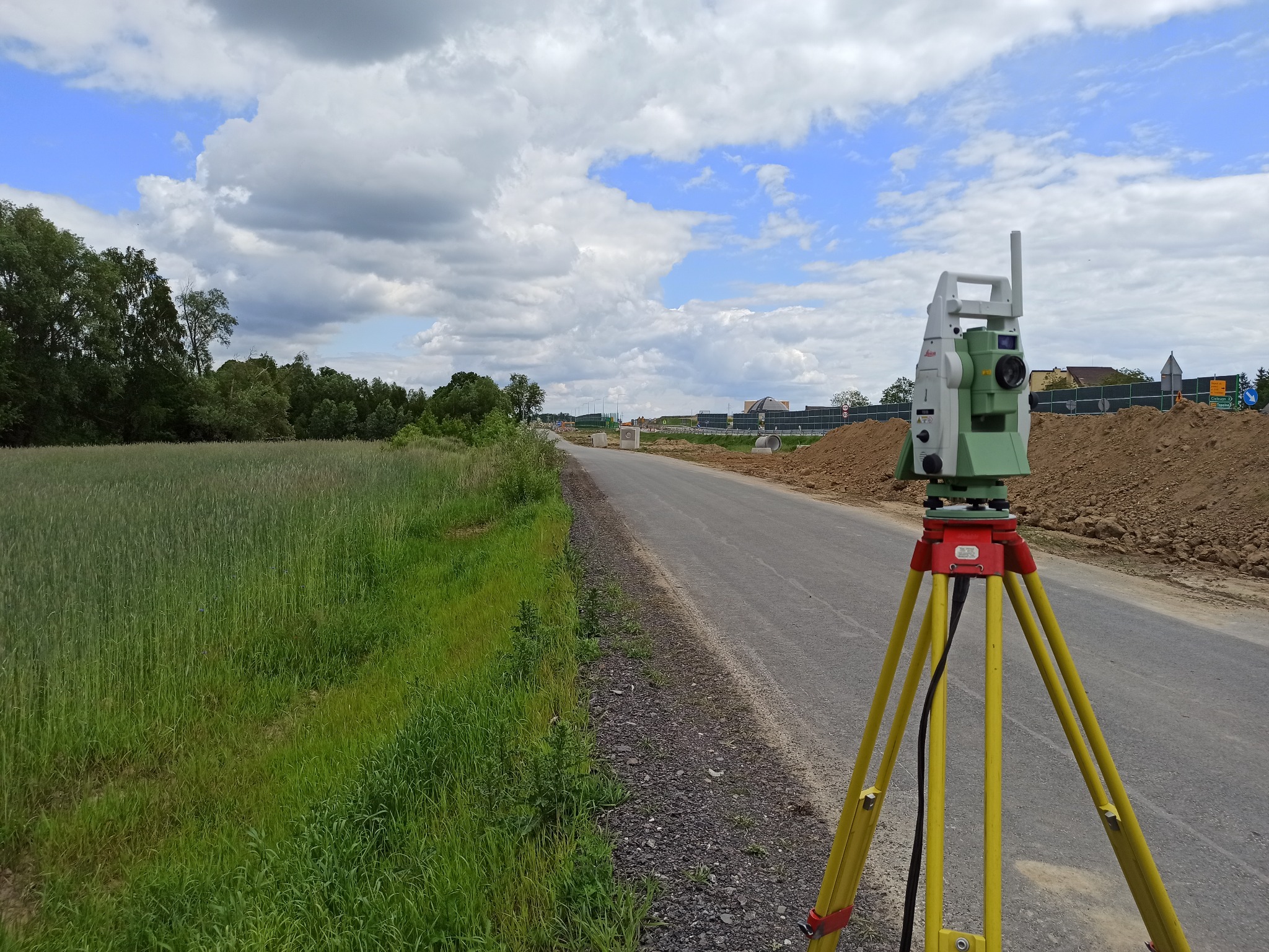 Tachimetr Leica na żółtym statywie przy nowej drodze w budowie, w tle pole i pochmurne niebo.