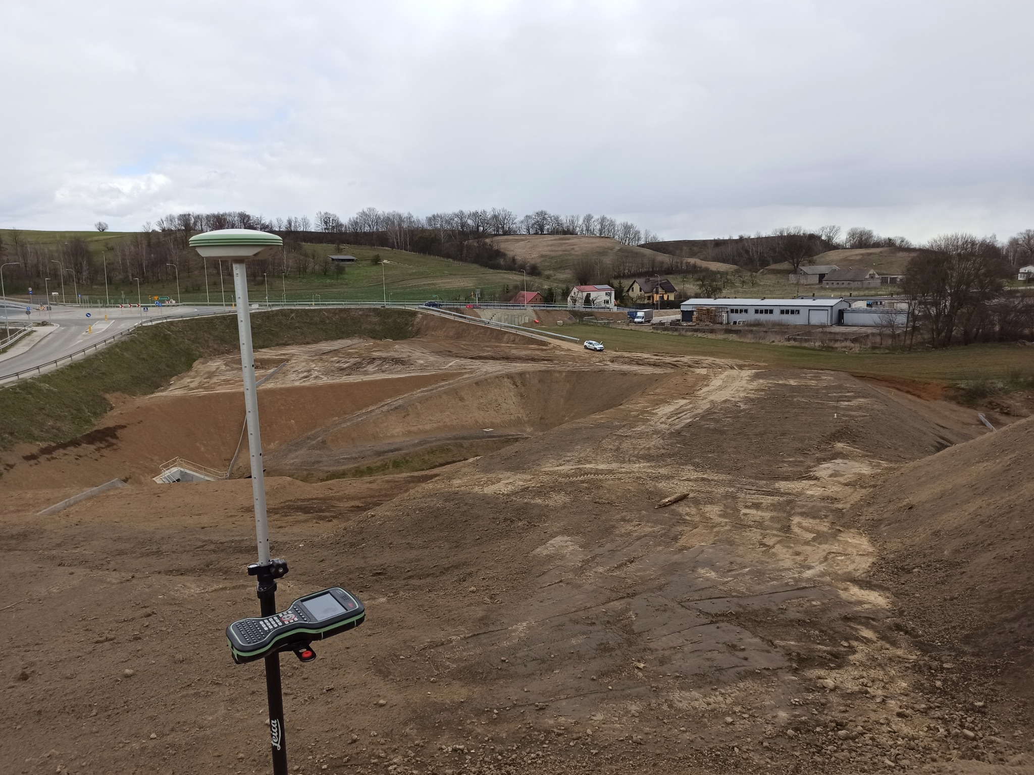 Pomiarowy odbiornik GNSS Leica na statywie, widok na wykop ziemny, w tle droga i zabudowania.