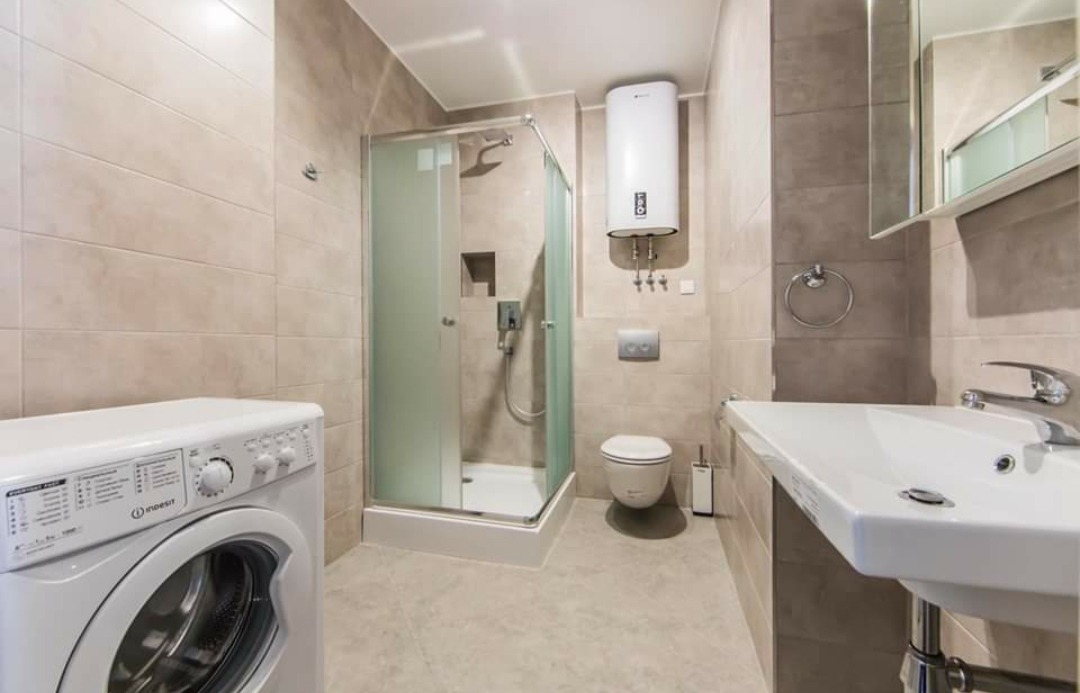 Jasna łazienka po remoncie z kabiną prysznicową, umywalką na postumencie, toaletą, bojlerem elektrycznym i pralką Indesit na pierwszym planie.