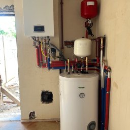 Gazownictwo-Ciepłownictwo-Hydraulika Jarosław Jurzysta - Nowoczesny, biały piec gazowy Termet z podłączeniami hydraulicznymi i zbiornikiem, zamontowany na surowej ścianie w trakcie instalacji. Widoczne rury i elementy instalacji.