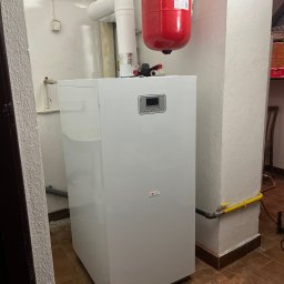 Gazownictwo-Ciepłownictwo-Hydraulika Jarosław Jurzysta - Biały, nowoczesny piec gazowy z czerwonym zbiornikiem wyrównawczym na tle ściany z instalacjami. Widoczna żółta rura gazowa i brązowa podłoga z terakoty.