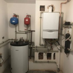 Biały piec gazowy Vaillant z instalacją, zbiornik wody i naczynia przeponowe w kotłowni. Widoczne rury miedziane i plastikowe. Ściany białe, podłoga szara.