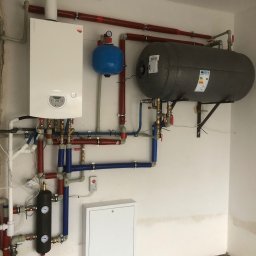 Biały piec gazowy Termet z szarym zasobnikiem wody i niebieskim naczyniem przeponowym, połączone rurami w kolorach czerwonym i niebieskim na tle białej ściany.