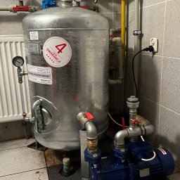 Hydrofor ze zbiornikiem hydroforowym Hydro-Vacuum, manometrami i niebieską pompą, zamontowany w pomieszczeniu z szarą glazurą. Widoczna instalacja wodna i elektryczna.