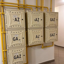 Zestaw sześciu beżowych szafek gazowych z żółtymi rurami, rozmieszczonych w dwóch rzędach na białej ścianie w korytarzu. Na szafkach widoczne numery i napisy 'GAZ'.