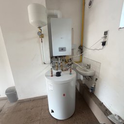 Gazownictwo-Ciepłownictwo-Hydraulika Jarosław Jurzysta - Biały kocioł gazowy i stojący zasobnik CWU z widocznym naczyniem wzbiorczym, żółtą rurą gazową oraz mosiężnymi zaworami i rurami instalacji grzewczej.