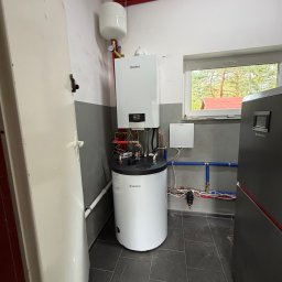 Gazownictwo-Ciepłownictwo-Hydraulika Jarosław Jurzysta - Kompletna instalacja grzewcza z białym kotłem gazowym Vaillant i cylindrycznym zasobnikiem c.w.u. w pomieszczeniu gospodarczym. Widoczne rury w izolacji, naczynie wzbiorcze i fragment ciemnoszarej...