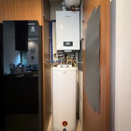 Gazownictwo-Ciepłownictwo-Hydraulika Jarosław Jurzysta - Biały kocioł gazowy Vaillant i zasobnik Galmet, z widocznymi rurami i zaworami, zamontowane w wąskiej drewnianej wnęce obok czarnej lodówki Samsung.