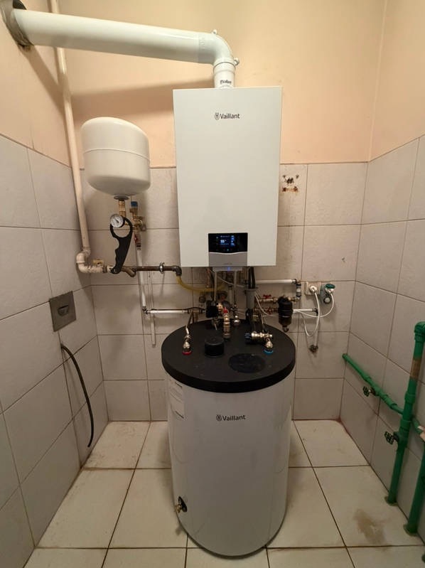 Biały, wiszący piec gazowy Vaillant z podłączonym zasobnikiem ciepłej wody w kotłowni z białymi płytkami. Widoczne rury i armatura.