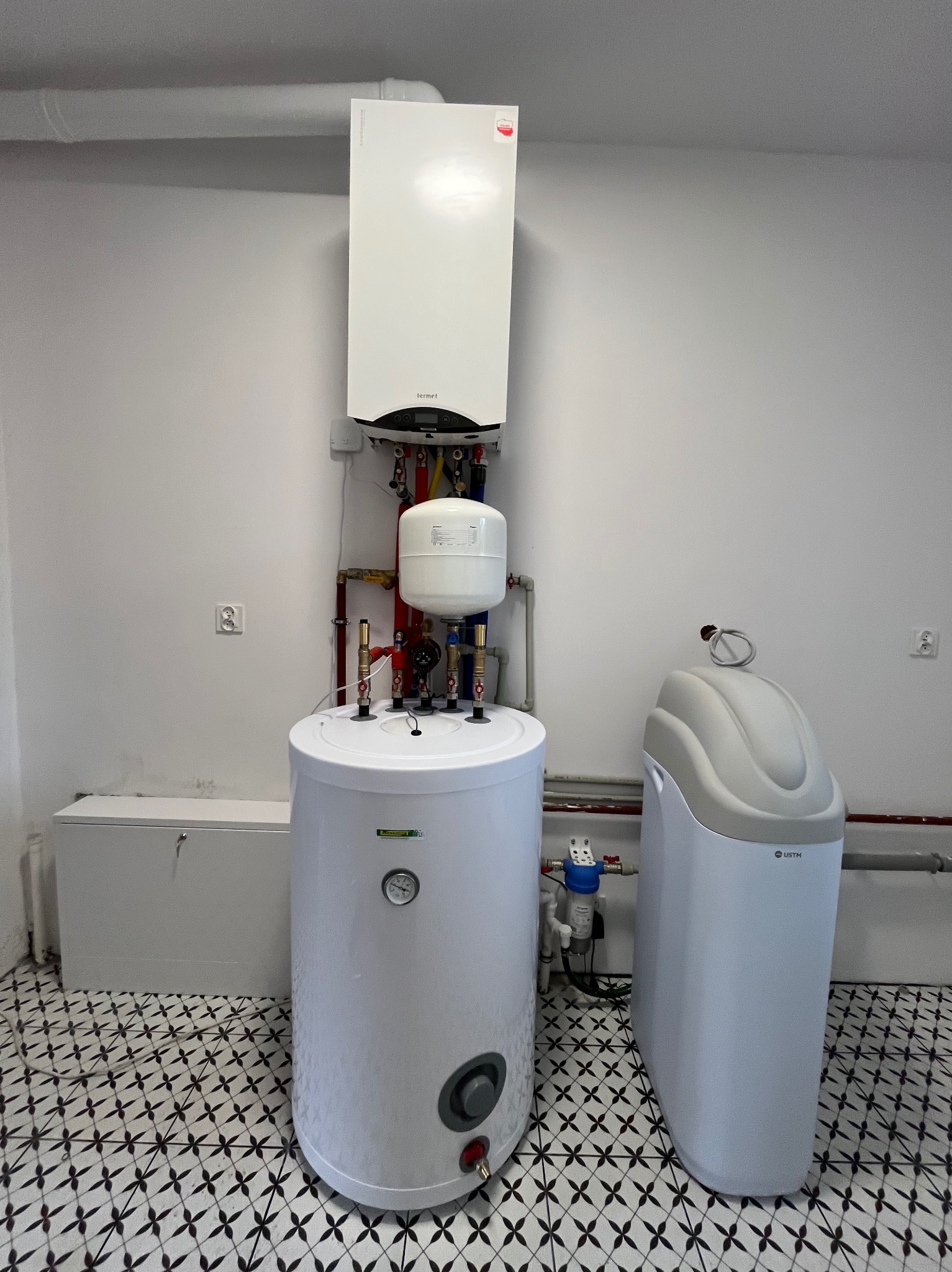 Biały piec gazowy wiszący nad bojlerem, obok zmiękczacz wody. Instalacja w pomieszczeniu z wzorzystą podłogą. Widoczne rury i zawory.