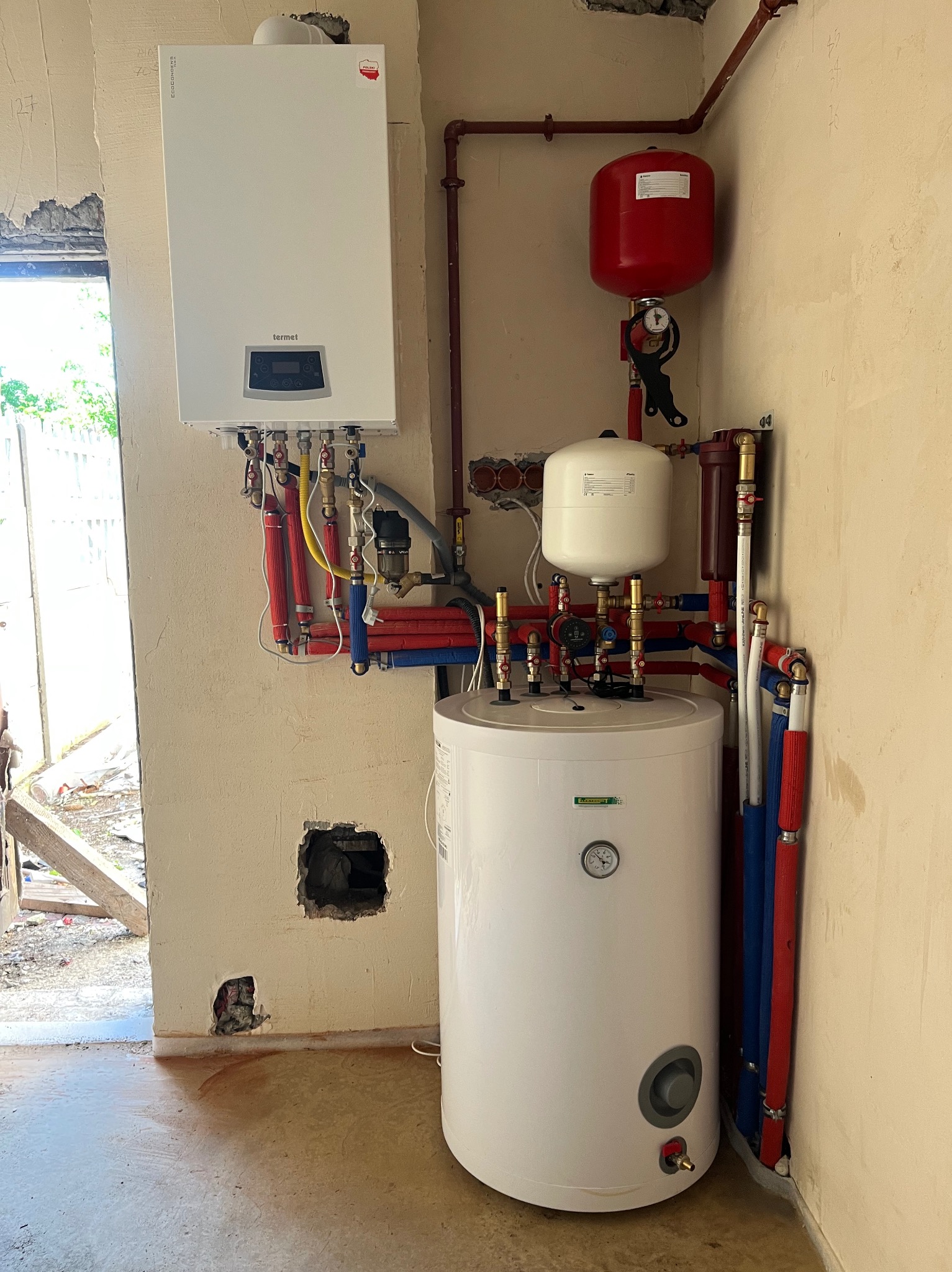 Nowoczesny, biały piec gazowy Termet z podłączeniami hydraulicznymi i zbiornikiem, zamontowany na surowej ścianie w trakcie instalacji. Widoczne rury i elementy instalacji.