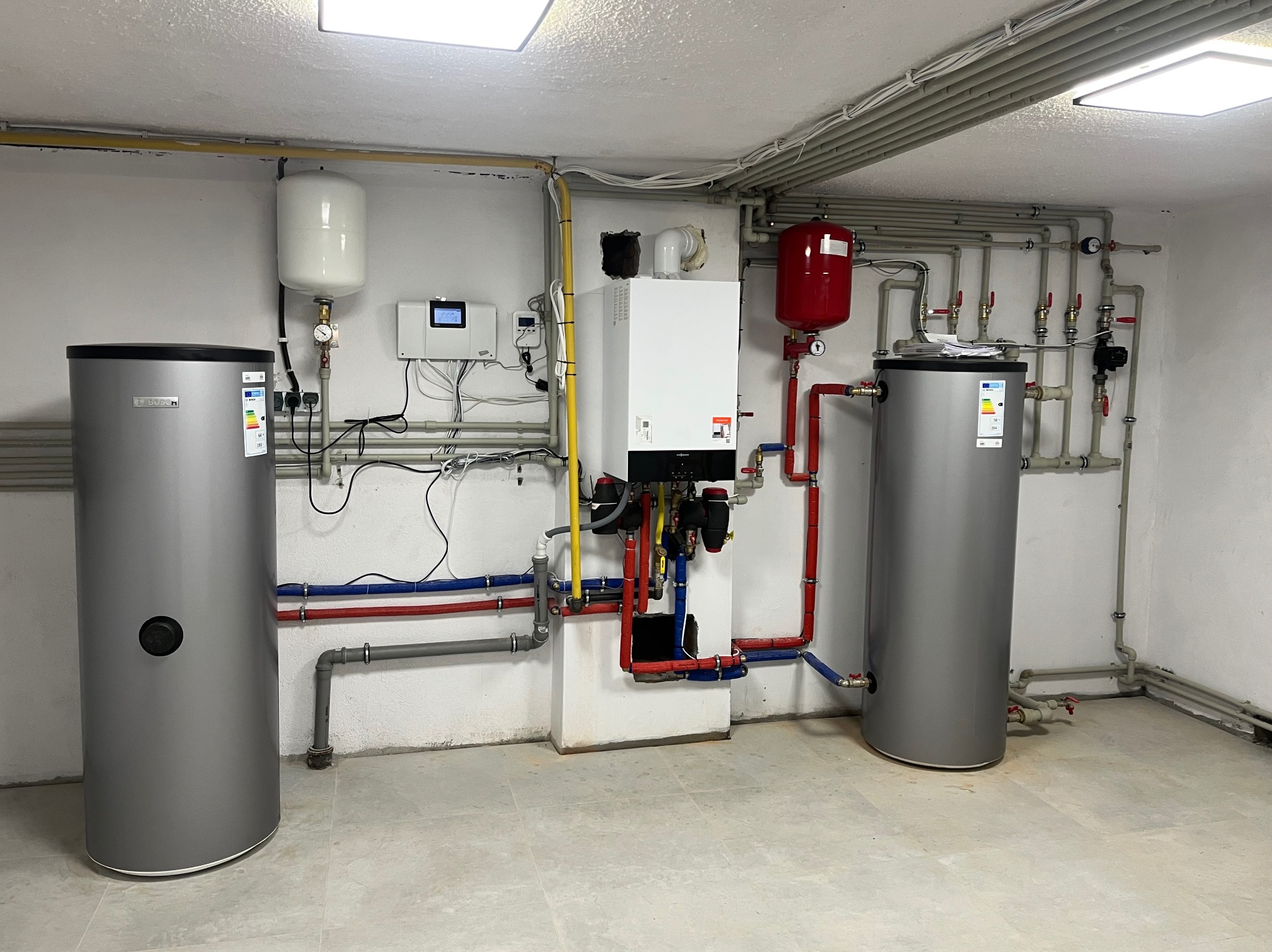 Nowoczesna kotłownia z białym piecem gazowym, dwoma szarymi zasobnikami ciepłej wody i rozbudowaną instalacją hydrauliczną z rurami w kolorach czerwonym i niebieskim.