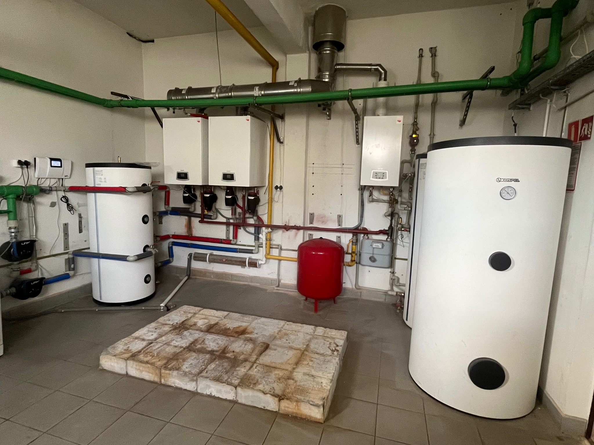 Kompletna kotłownia z dwoma piecami, zbiornikiem Kospel, czerwonym zbiornikiem wyrównawczym i rozbudowaną instalacją rur w kolorach: zielonym, czerwonym, niebieskim i żółtym.