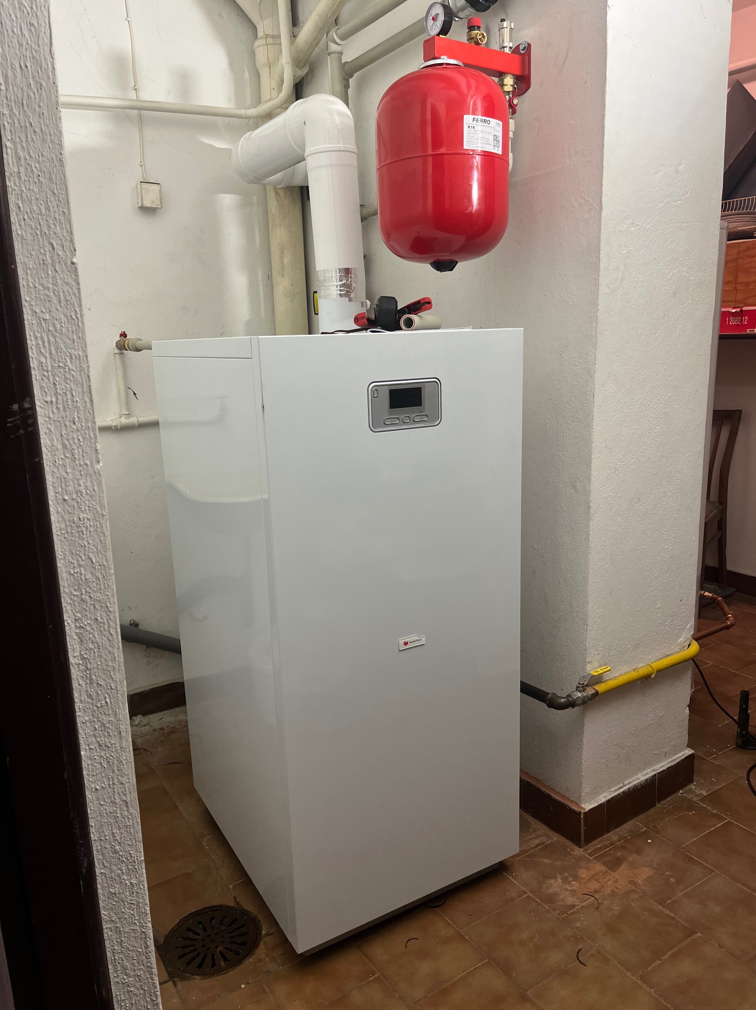 Biały, nowoczesny piec gazowy z czerwonym zbiornikiem wyrównawczym na tle ściany z instalacjami. Widoczna żółta rura gazowa i brązowa podłoga z terakoty.