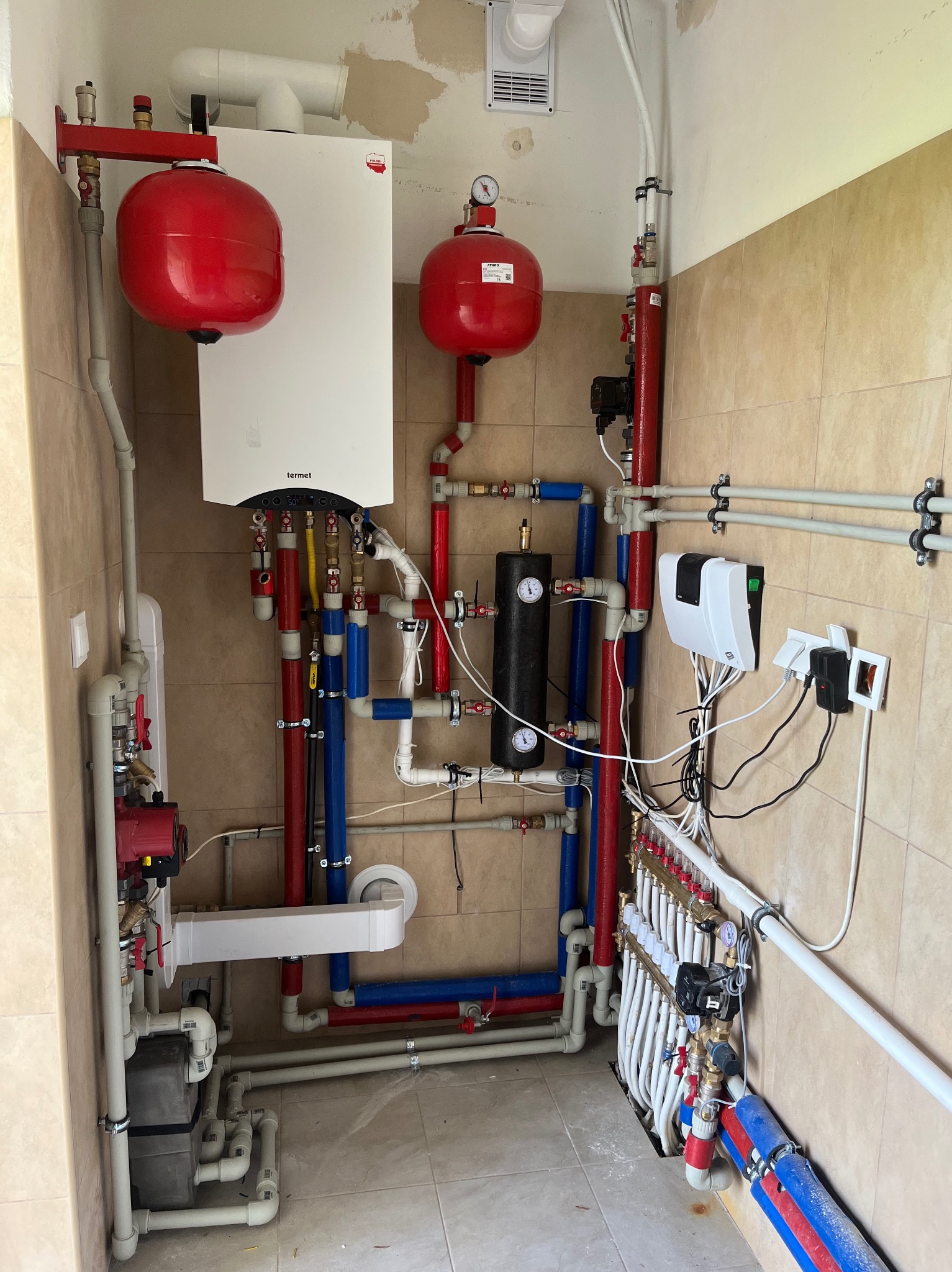 Kompletna instalacja grzewcza z piecem Termet, zbiornikami wyrównawczymi i rozdzielaczem podłogówki. Rury w kolorach czerwonym i niebieskim. Ściany wyłożone płytkami.