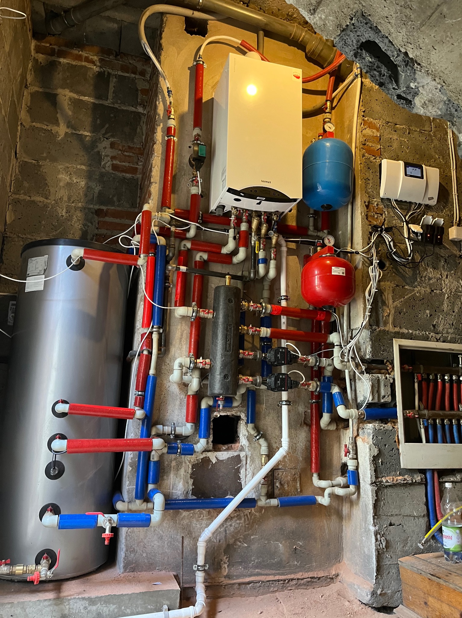 Złożona instalacja grzewcza z kotłem gazowym Termet, zbiornikiem i rurami w kolorach czerwonym i niebieskim, zamontowana na tle surowej, ceglanej ściany w piwnicy.