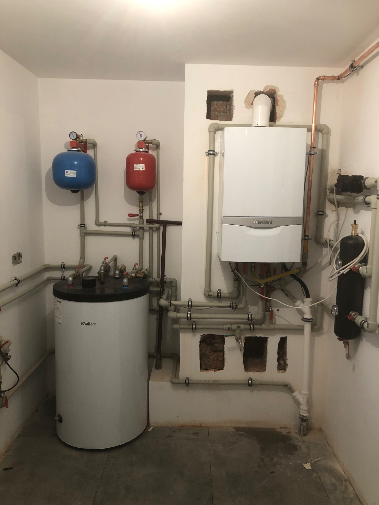 Biały piec gazowy Vaillant z instalacją, zbiornik wody i naczynia przeponowe w kotłowni. Widoczne rury miedziane i plastikowe. Ściany białe, podłoga szara.