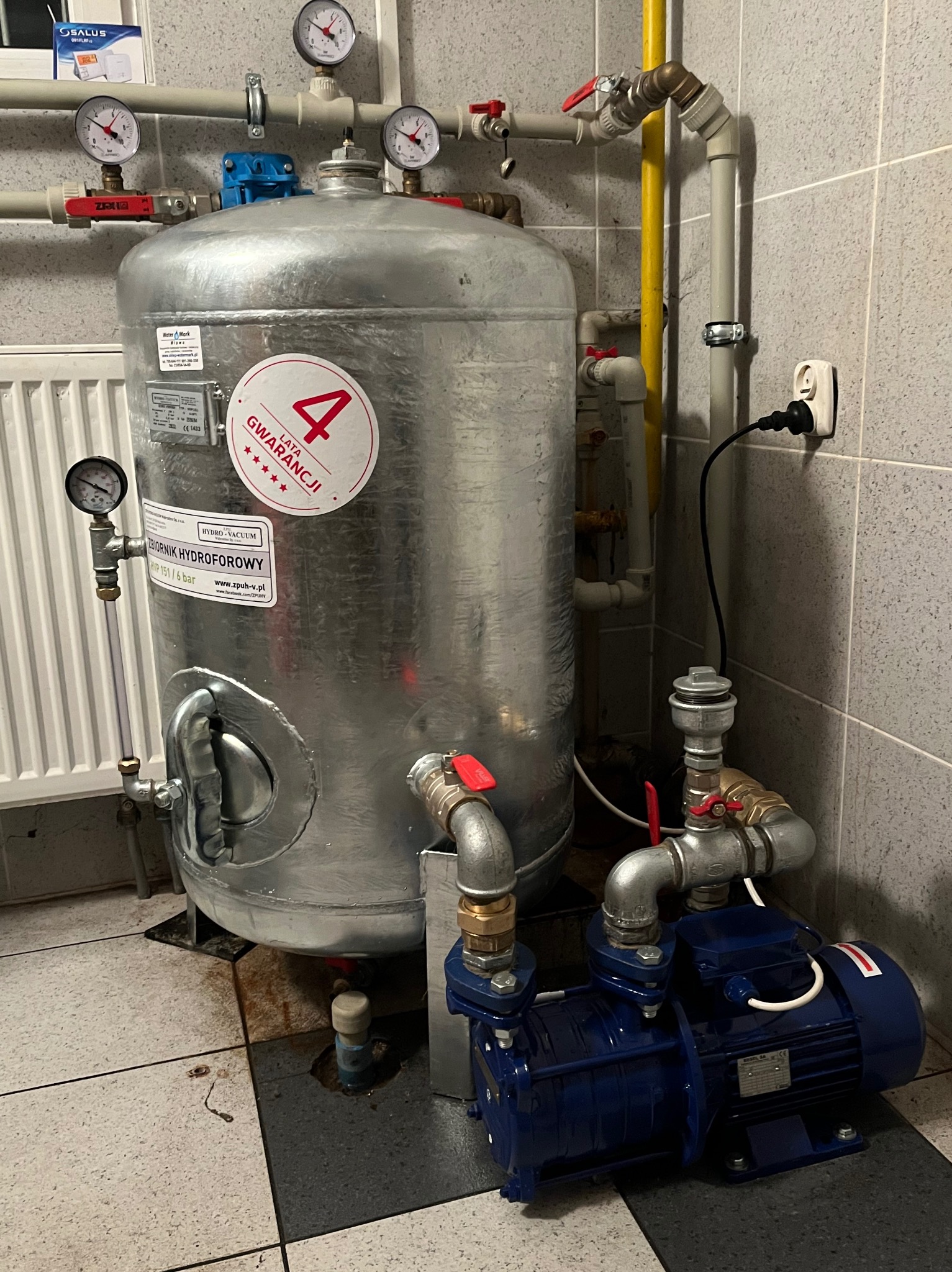 Hydrofor ze zbiornikiem hydroforowym Hydro-Vacuum, manometrami i niebieską pompą, zamontowany w pomieszczeniu z szarą glazurą. Widoczna instalacja wodna i elektryczna.