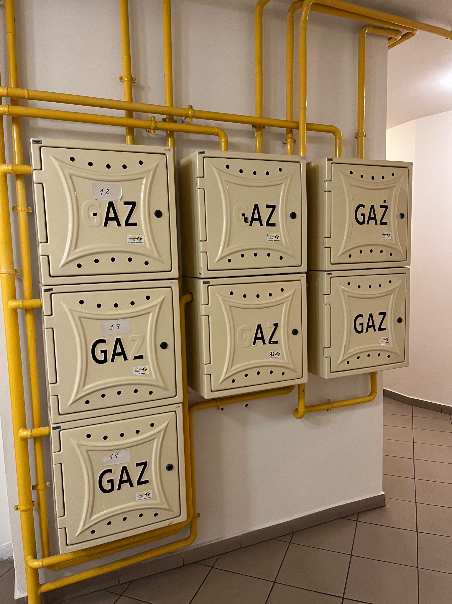 Zestaw sześciu beżowych szafek gazowych z żółtymi rurami, rozmieszczonych w dwóch rzędach na białej ścianie w korytarzu. Na szafkach widoczne numery i napisy 'GAZ'.