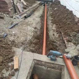 Wykop z pomarańczową rurą kanalizacyjną ułożoną w ziemi, widoczna studzienka rewizyjna z wchodzącą do niej rurą karbowaną, w tle mur z bloczk&oacute;w betonowych i biała ściana, obok leżą płytki...