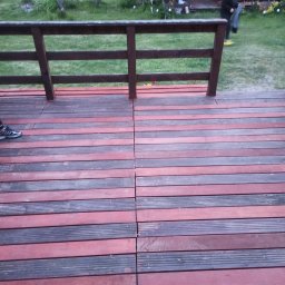 Andrzej Wojciechowski - Dwukolorowy taras z desek kompozytowych, widok z góry, z fragmentem buta sportowego w lewym dolnym rogu i drewnianą balustradą w tle, za którą widać altanę i osobę w żółtych butach.