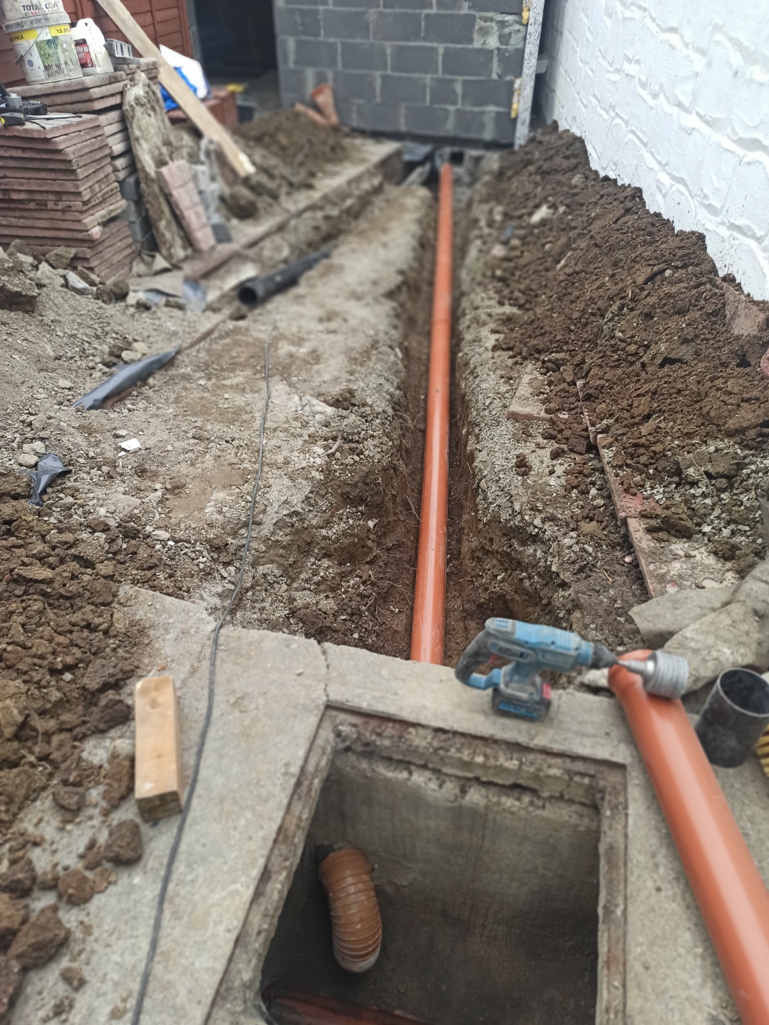 Wykop z pomarańczową rurą kanalizacyjną ułożoną w ziemi, widoczna studzienka rewizyjna z wchodzącą do niej rurą karbowaną, w tle mur z bloczków betonowych i biała ściana, obok leżą płytki...