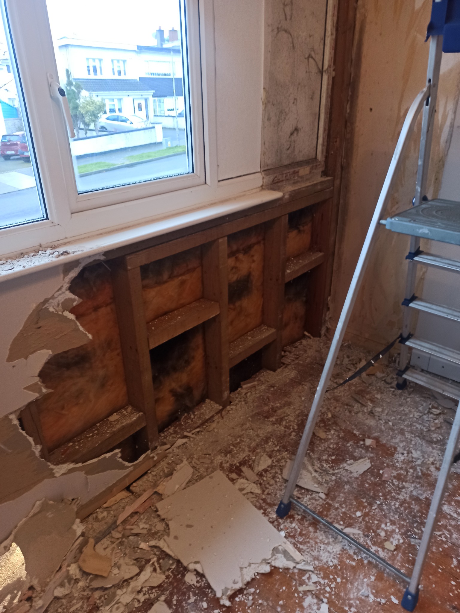 Fragment ściany z odsłoniętą izolacją z wełny mineralnej i drewnianą konstrukcją pod oknem, częściowo zerwana warstwa wierzchnia ściany, obok oparta drabina aluminiowa, widoczne ślady gruzu...