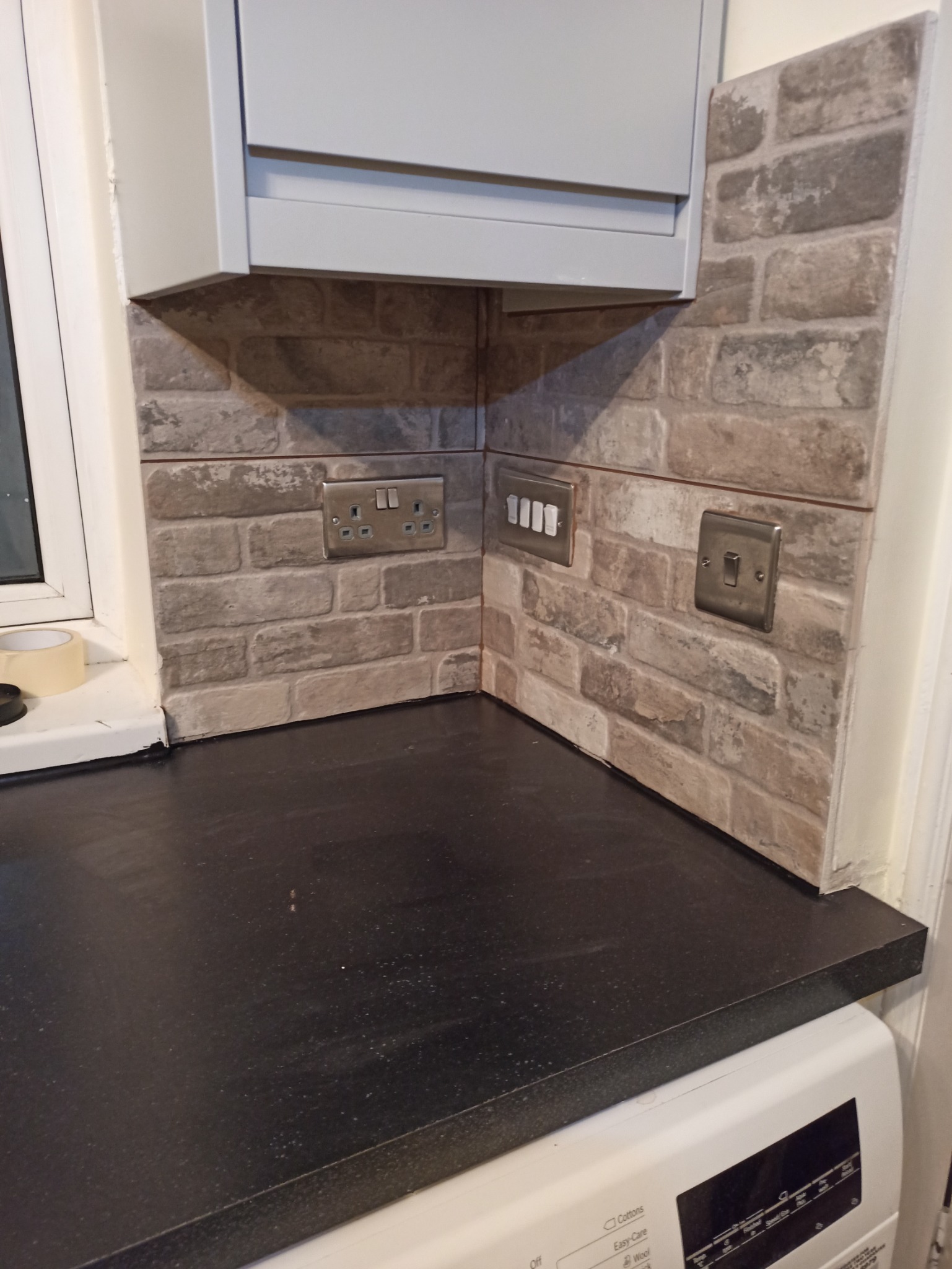 Wykończona ściana nad blatem kuchennym z płytkami imitującymi cegłę w odcieniach szarości i beżu, z zamontowanymi gniazdkami i włącznikami w metalowych ramkach, pod szafką kuchenną.