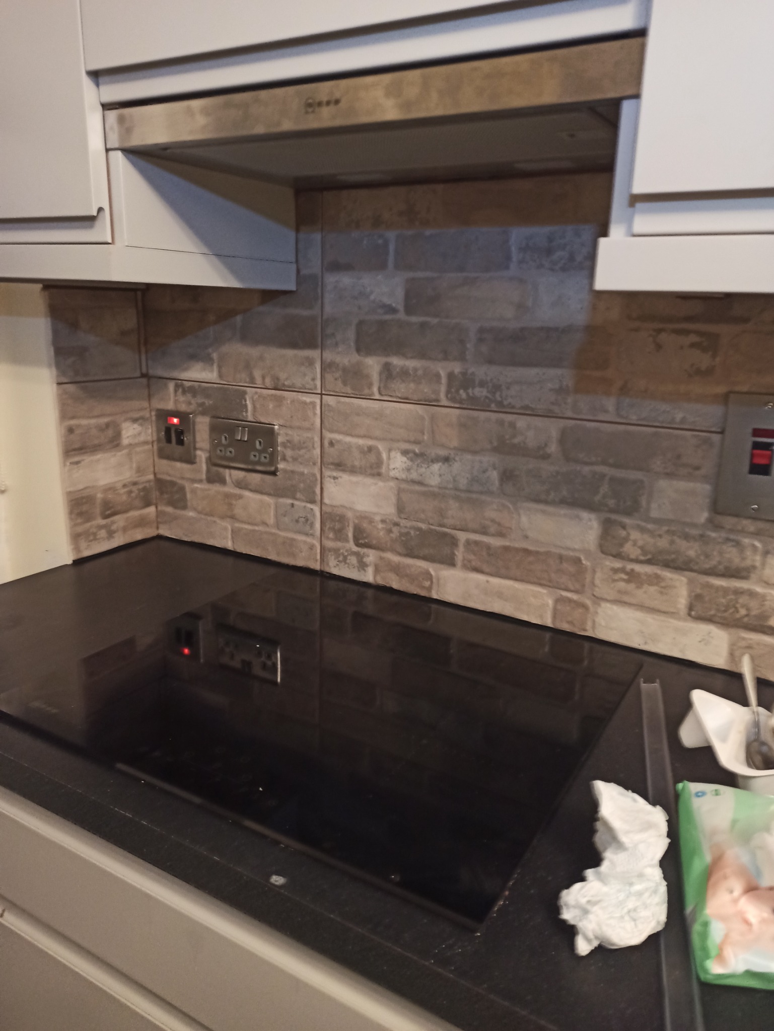 Kuchnia z płytą indukcyjną, okładziną ścienną imitującą cegłę, szarymi szafkami i okapem, widoczne gniazdka elektryczne.