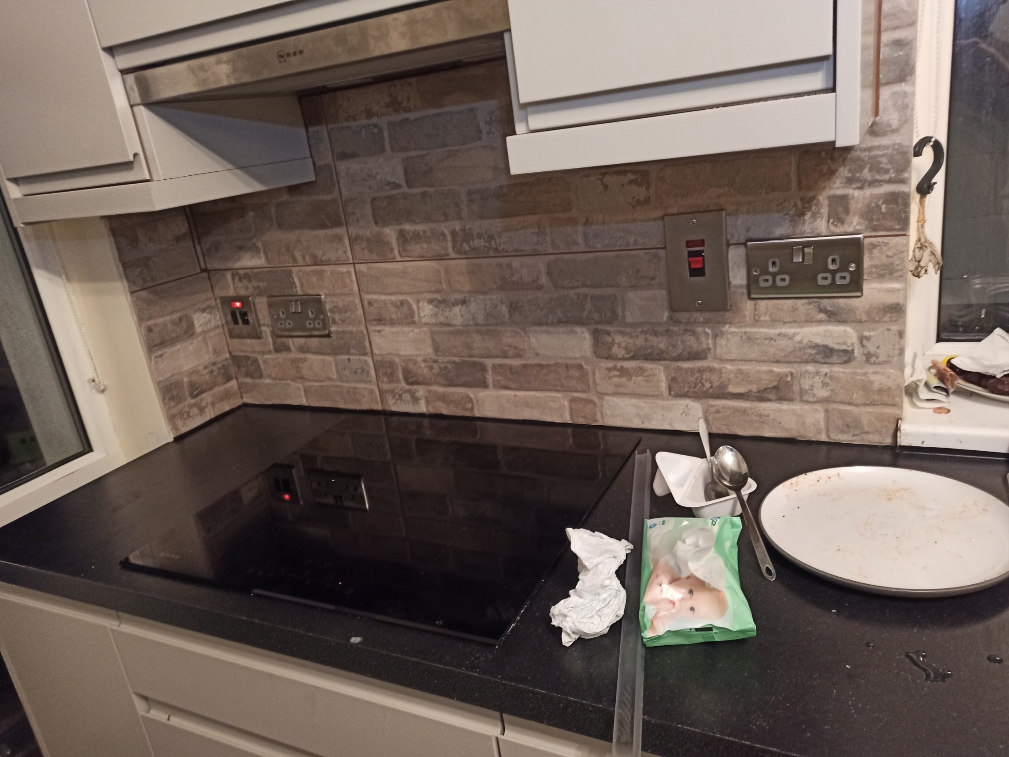Kuchnia po remoncie: ściana z płytek imitujących cegłę, czarny blat z płytą indukcyjną, widoczne gniazdka elektryczne i włączniki, resztki po posiłku na blacie.