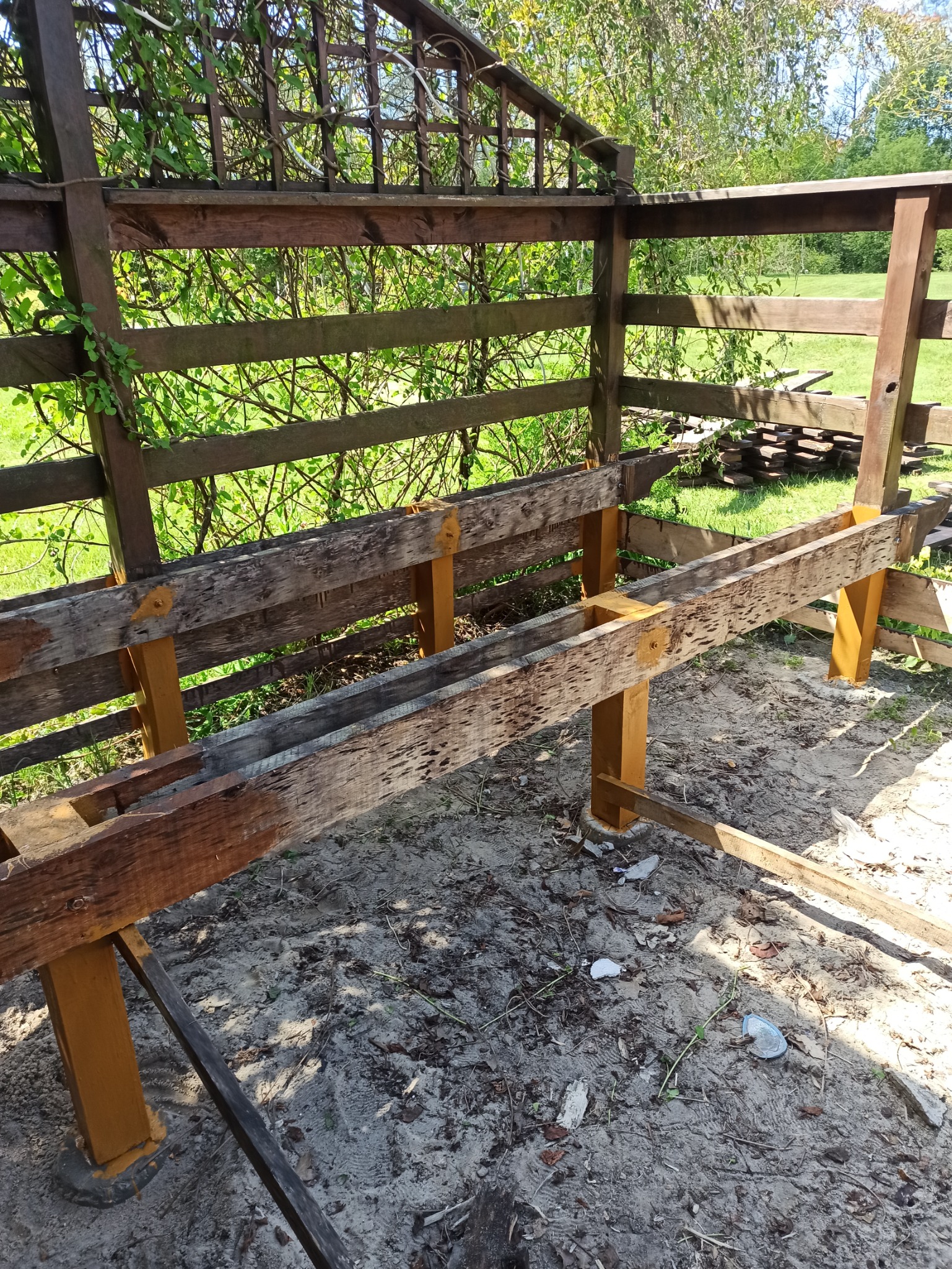 Konstrukcja drewniana z belek i słupków pomalowanych na żółto, tworząca fragment pergoli lub altany w trakcie budowy, widoczne elementy kratownicy i roślinność w tle.