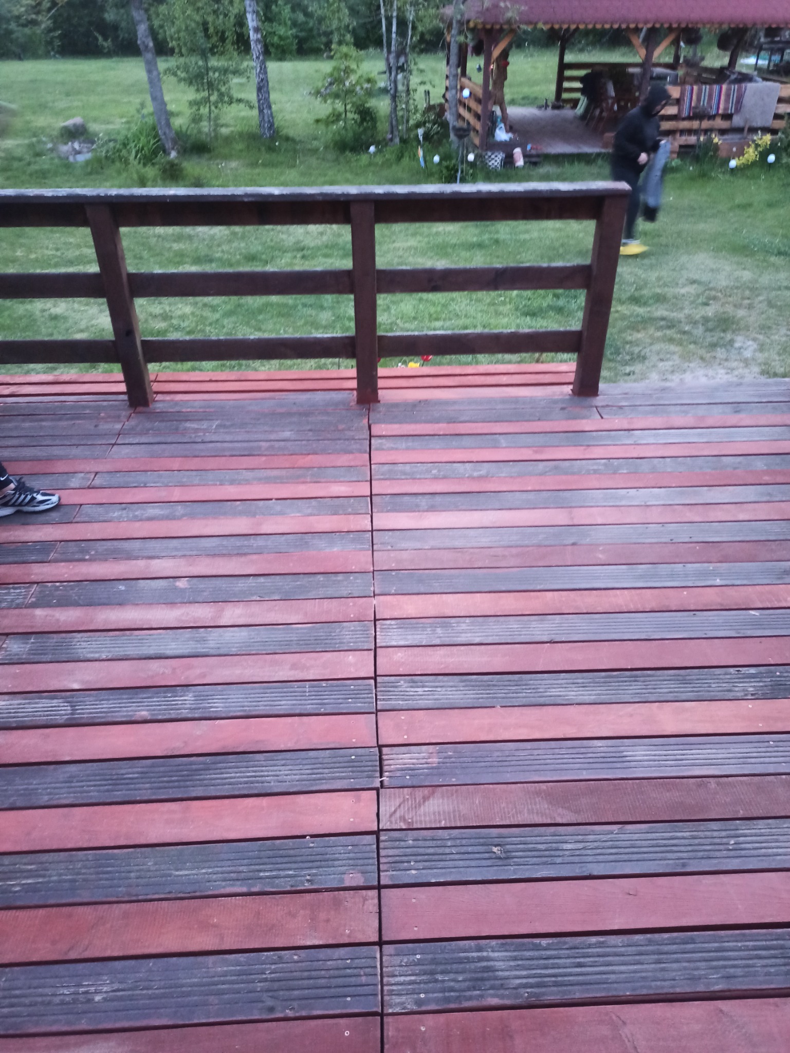 Dwukolorowy taras z desek kompozytowych, widok z góry, z fragmentem buta sportowego w lewym dolnym rogu i drewnianą balustradą w tle, za którą widać altanę i osobę w żółtych butach.