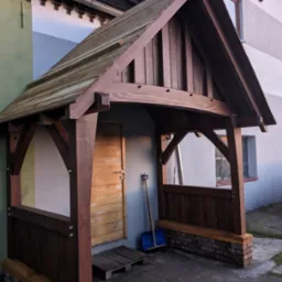 Drewniany daszek nad wejściem z drewnianymi ściankami bocznymi na ceglanej podmurówce, niebieska łopata oparta o ścianę, drewniane drzwi wejściowe z poziomymi deskami.