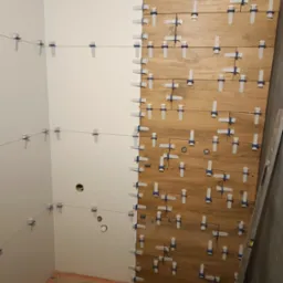 Proces układania drewnopodobnych płytek w łazience, z użyciem systemu poziomowania płytek na ścianie i sznurka wyznaczającego linię na sąsiedniej ścianie, widoczne otwory na instalacje.