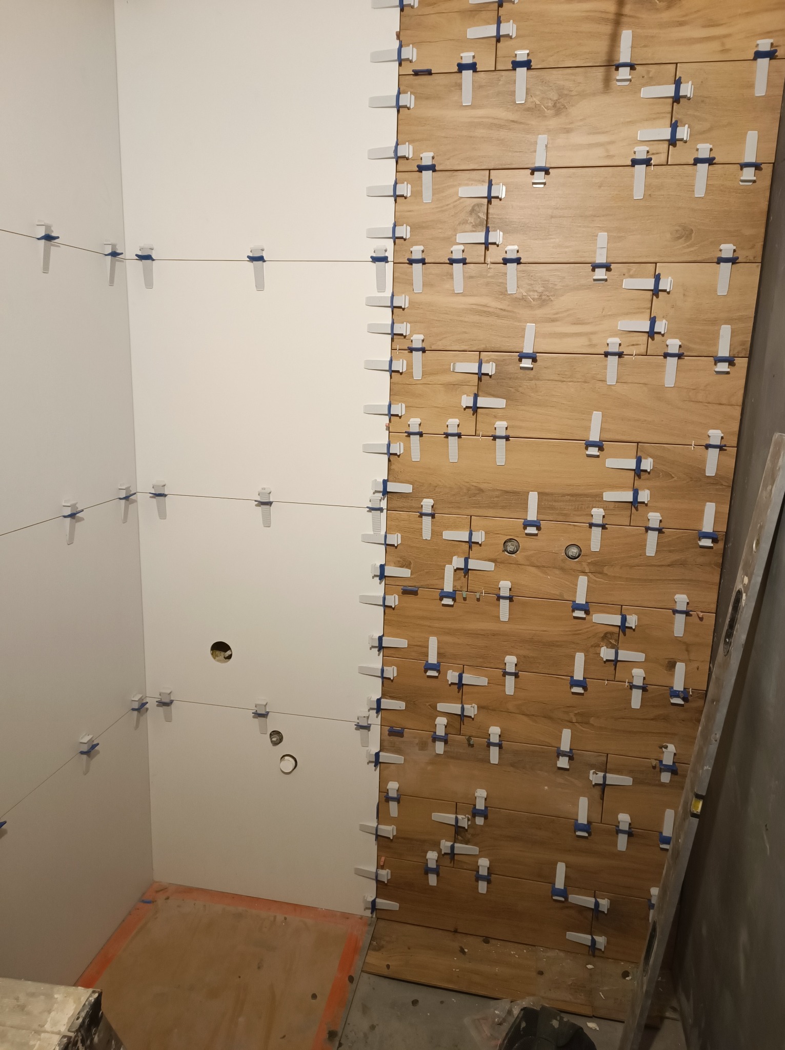 Proces układania drewnopodobnych płytek w łazience, z użyciem systemu poziomowania płytek na ścianie i sznurka wyznaczającego linię na sąsiedniej ścianie, widoczne otwory na instalacje.