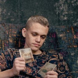 Wilku photography - Młody mężczyzna siedzi na wzorzystej kanapie, trzymając w dłoniach banknoty o niskim nominale i spoglądając na nie zamyślonym wzrokiem, ubrany w koszulę w paisley.