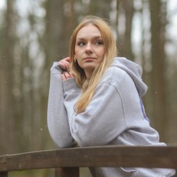 Wilku photography - Portret kobiety w szarej bluzie z kapturem, opierającej się o drewnianą barierkę na tle zamazanego lasu w zimowej scenerii.