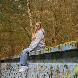 Wilku photography - Młoda kobieta w bluzie z kapturem i dżinsach siedzi na pomalowanej na niebiesko, pokrytej mchem betonowej konstrukcji na tle drzew bez liści.