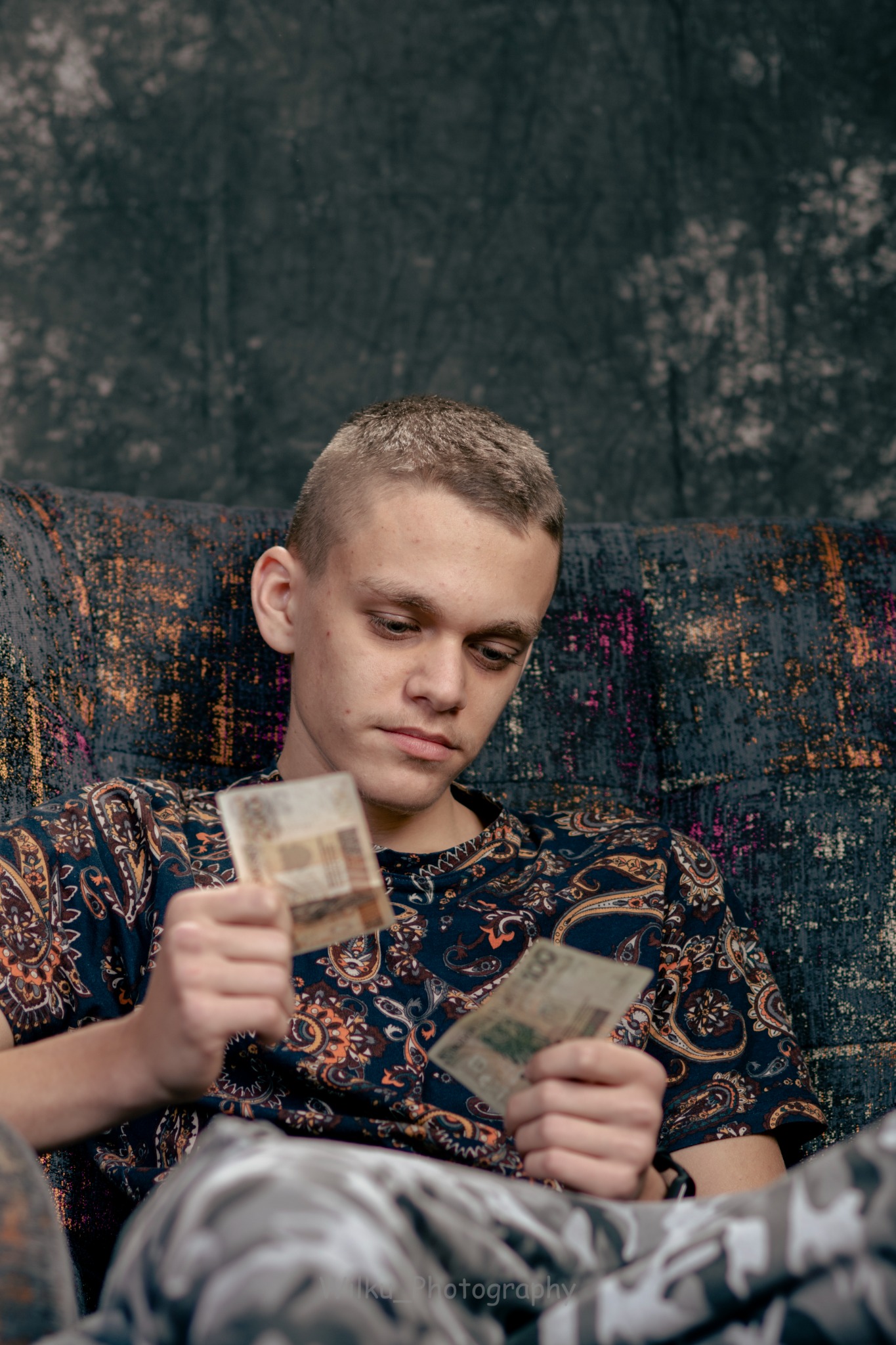 Młody mężczyzna siedzi na wzorzystej kanapie, trzymając w dłoniach banknoty o niskim nominale i spoglądając na nie zamyślonym wzrokiem, ubrany w koszulę w paisley.