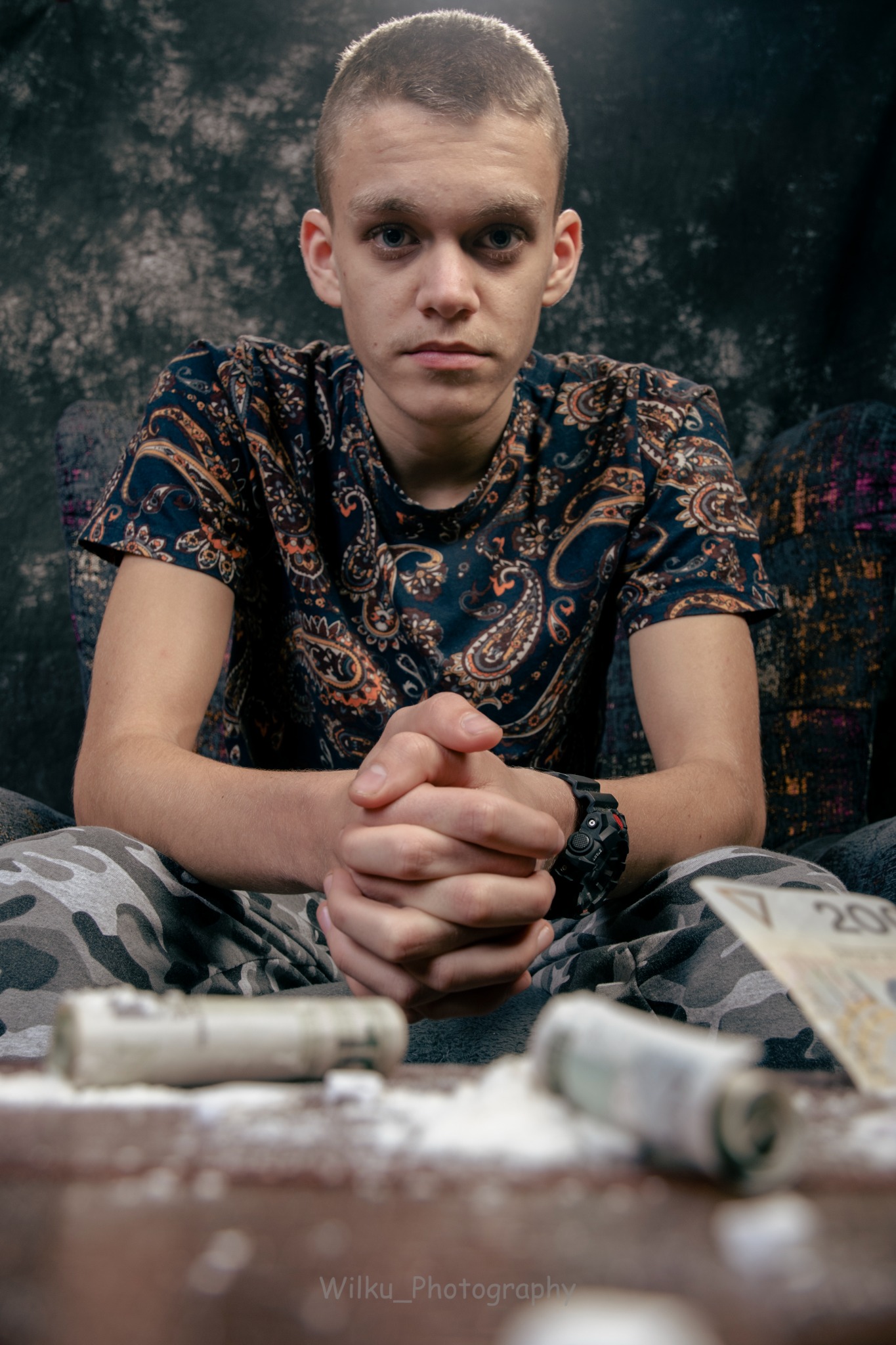 Młody mężczyzna z poważnym wyrazem twarzy siedzi na fotelu z złożonymi dłońmi, na stole przed nim zwinięte banknoty i biały proszek.