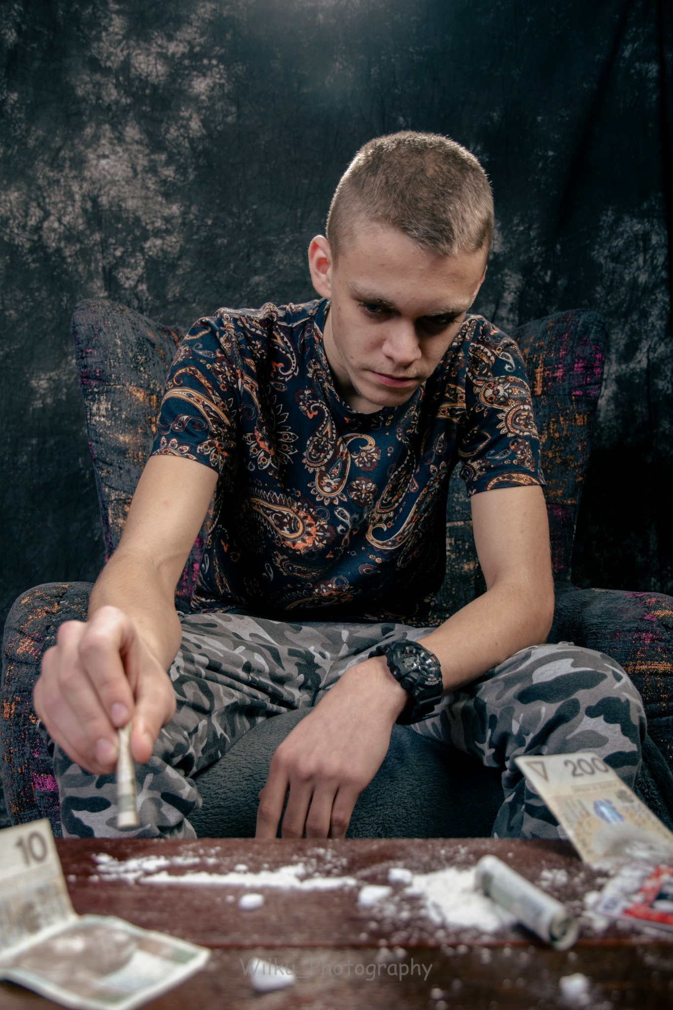 Młody mężczyzna siedzi w fotelu, pochylony nad stołem z rozsypanym białym proszkiem, zwiniętym banknotem i pieniędzmi, ubrany w koszulkę w paisley i spodnie moro, z czarnym zegarkiem na ręku.