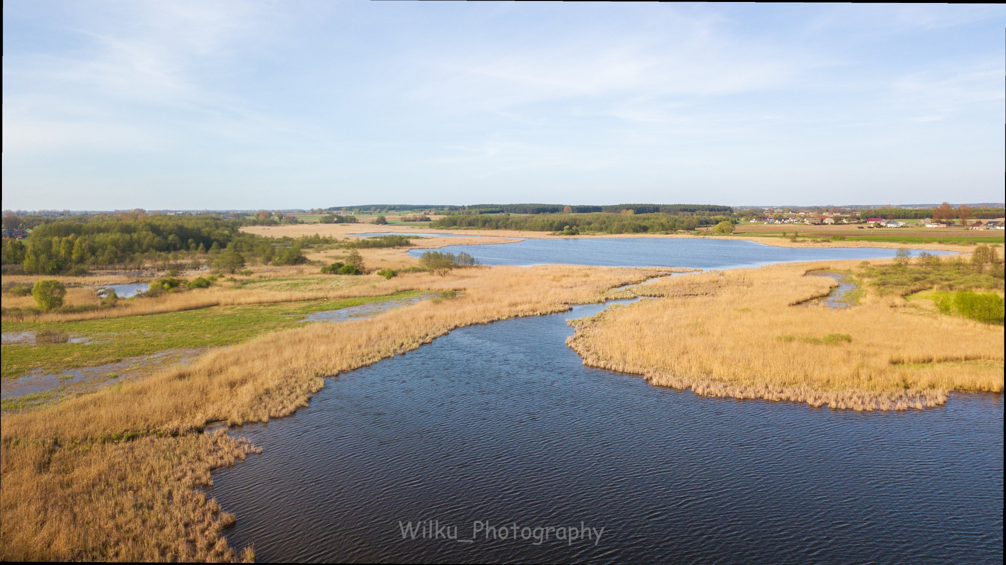 Panoramiczny widok z lotu ptaka na rozległe jezioro otoczone złotymi trzcinami i zielonymi drzewami, z oddalonymi zabudowaniami w tle pod jasnym, błękitnym niebem.