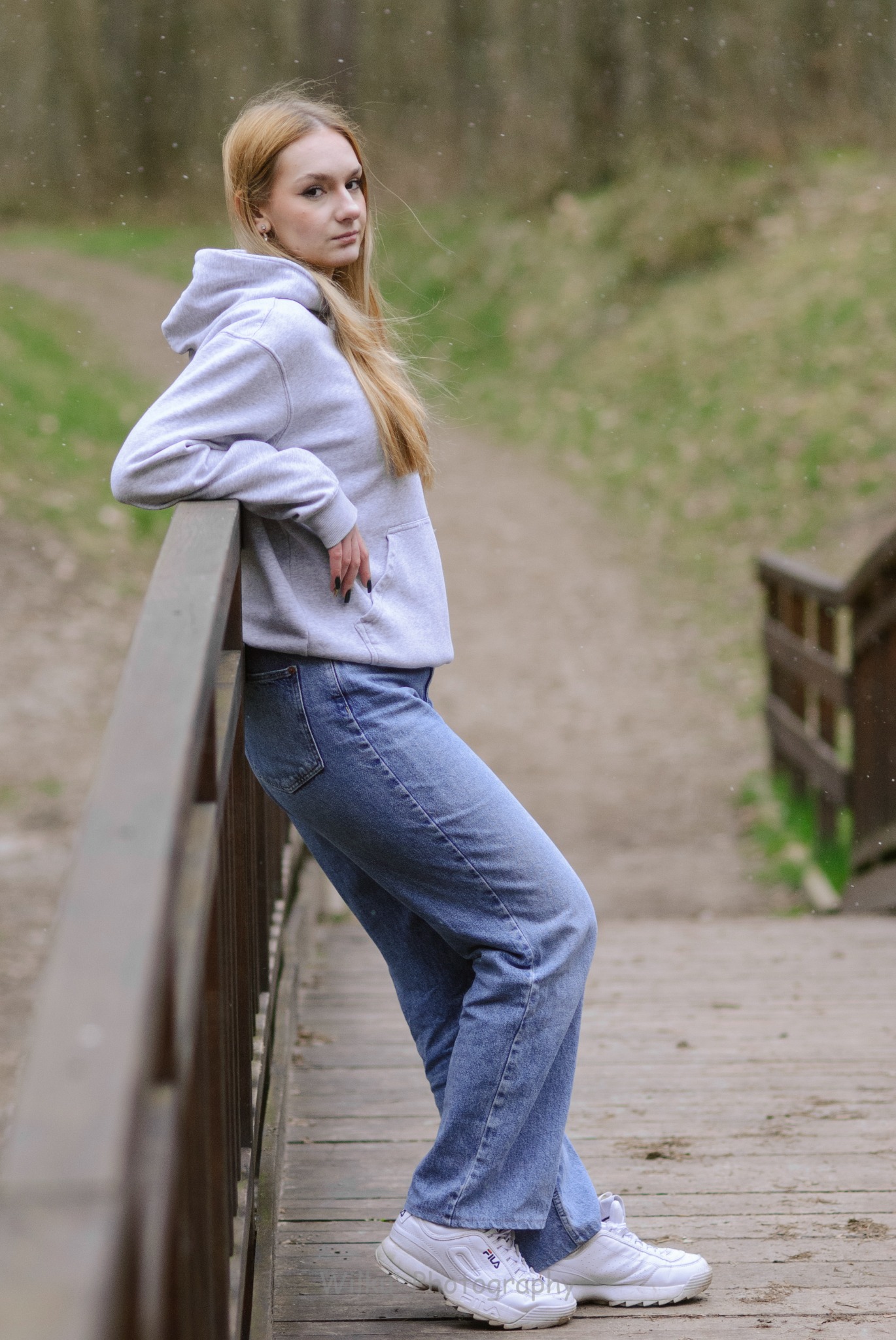 Portret kobiety w bluzie z kapturem i jeansach, opierającej się o drewnianą barierkę mostu w parku, z lekkim uśmiechem i spojrzeniem w obiektyw.