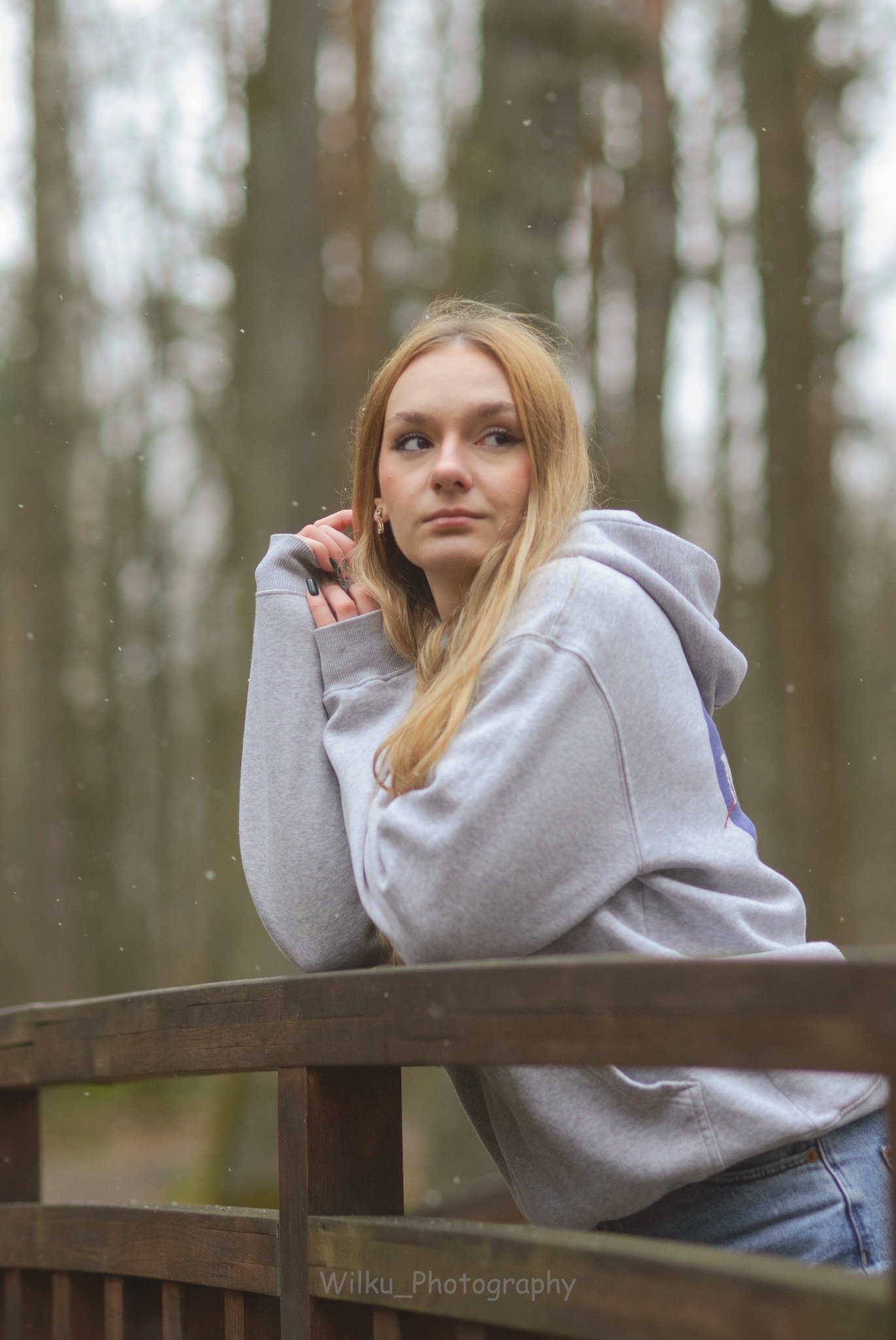 Portret kobiety w szarej bluzie z kapturem, opierającej się o drewnianą barierkę na tle zamazanego lasu w zimowej scenerii.