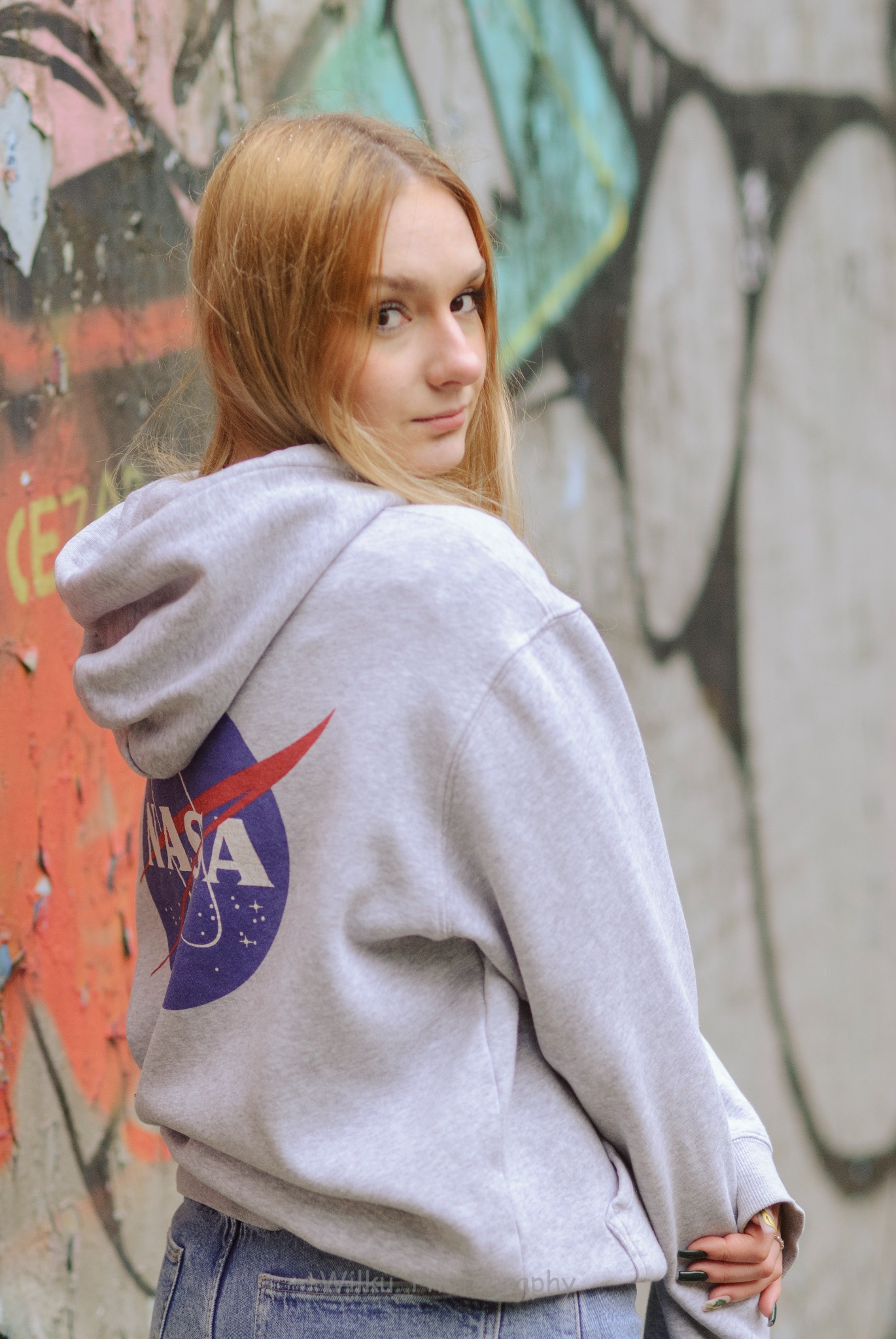 Młoda kobieta w szarej bluzie z kapturem z logo NASA, odwrócona przez ramię, na tle ściany pokrytej graffiti.